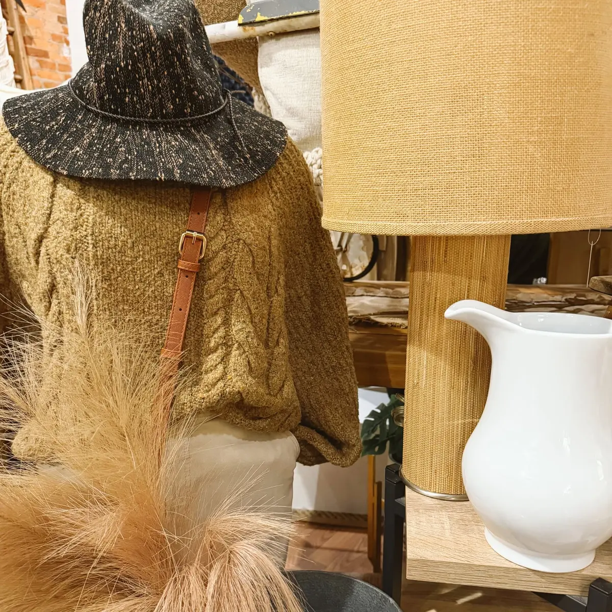 Olive cable-knit sweater and speckled wide-brim hat beside a woven lamp and white pitcher
